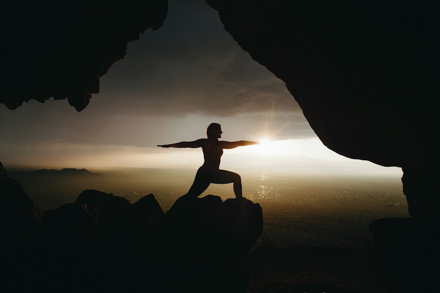 Silhouette of a yoga practitioner holding warrior pose beneath a coastal rock arch at sunrise